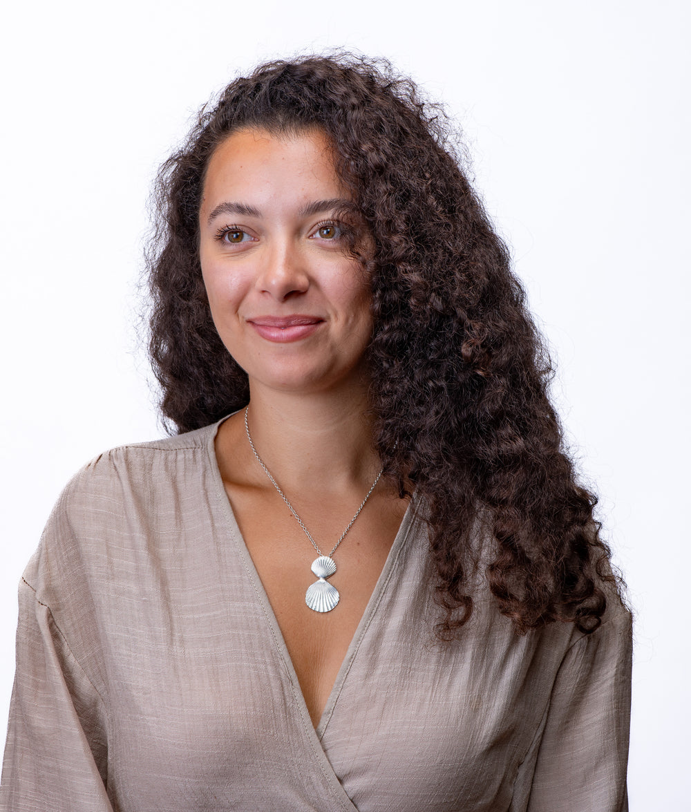 Woman with long, curly hair wearing a beige top and double scallop shell necklace on a white background