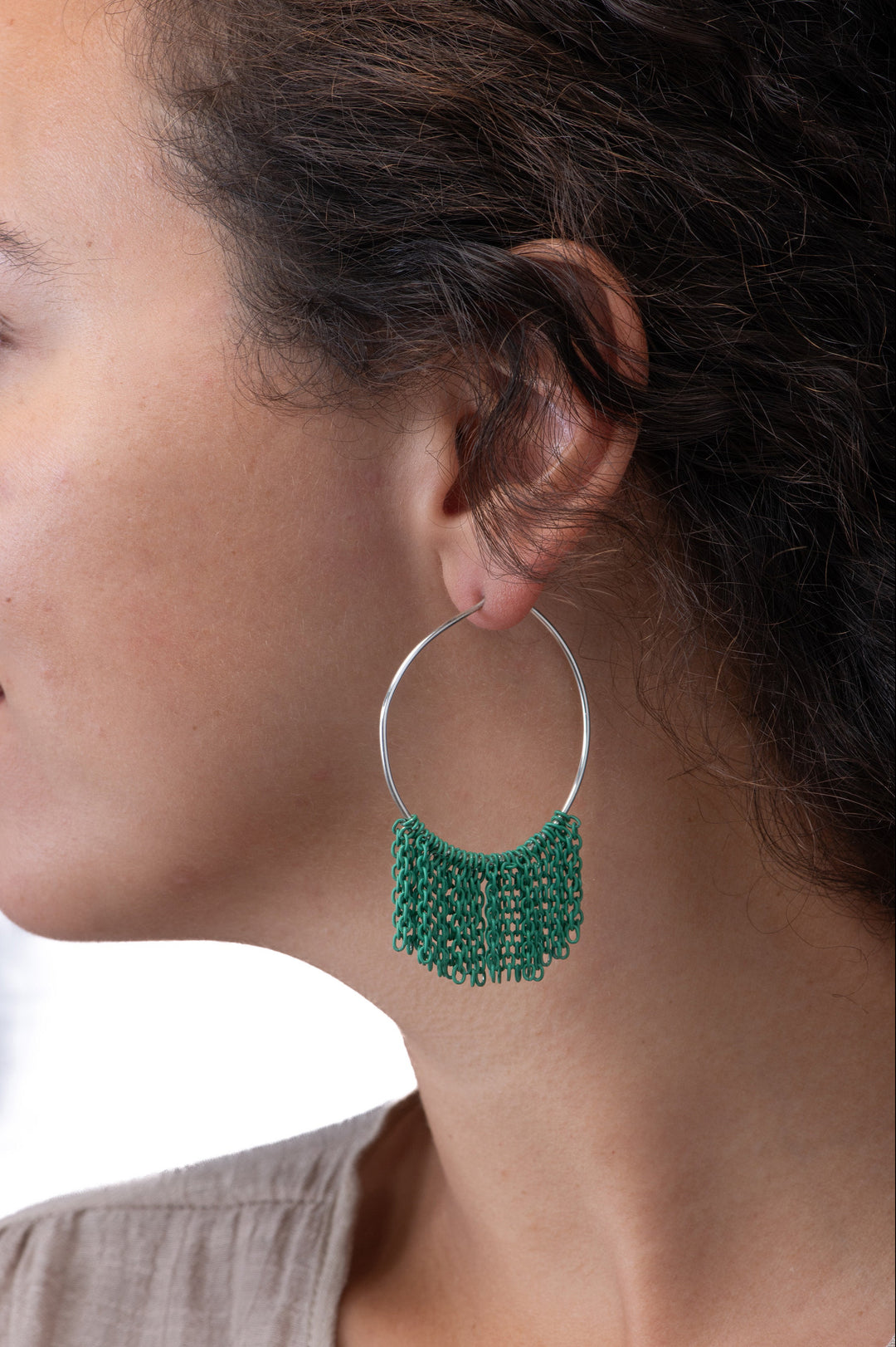 Close-up of a person wearing silver hoop earrings with green fringe chain on a neutral background