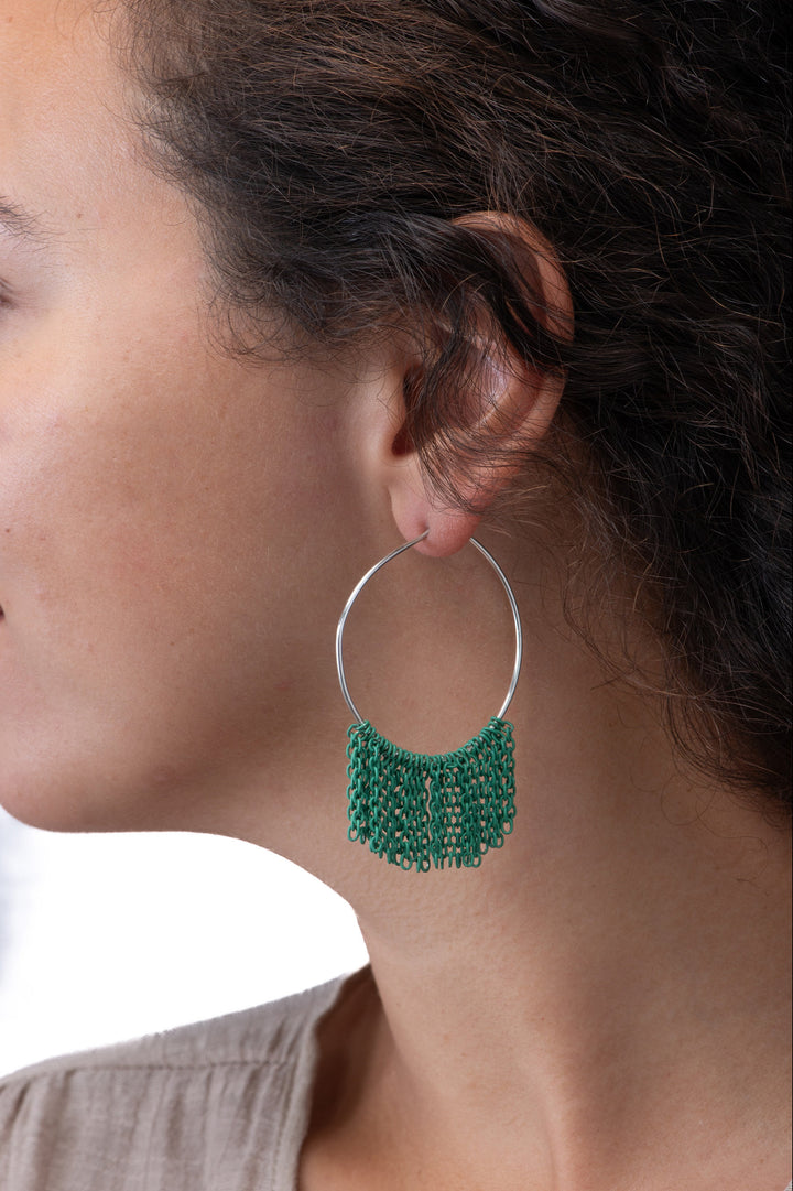 Close-up of a person wearing silver hoop earrings with green fringe chain on a neutral background