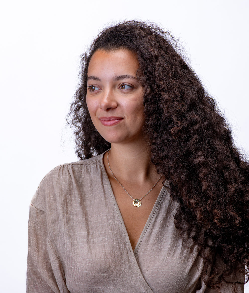 Woman with long, curly hair wearing a beige top against a white background with two tone gold and silver necklace.